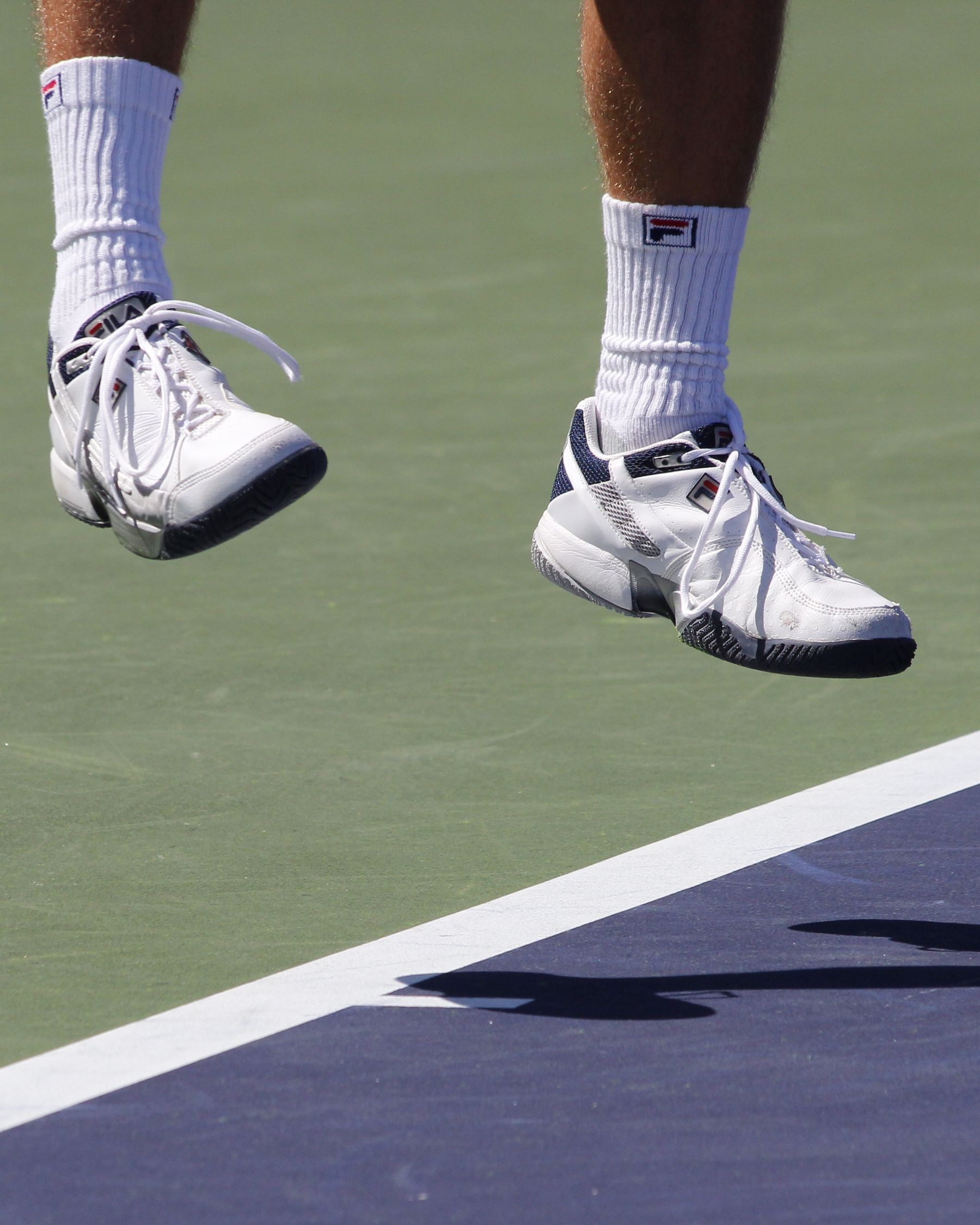 Close-up of an adult tennis player’s feet airborne above a hard court during footwork.